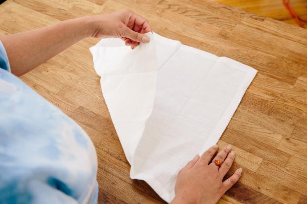 Place a square napkin on a table with the corners pointing at north and south directions. Fold the napkin over itself to make a triangle shape. Keep folding the napkin over itself to create smaller and smaller triangle shapes. Then wrap two rubber bands around the tightly folded triangle napkin.