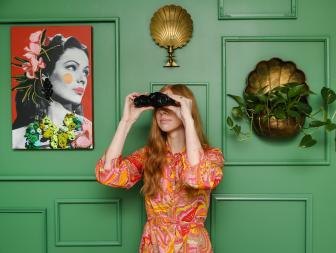 Woman Stands In Front of Green Gallery Wall Holding Up Binoculars