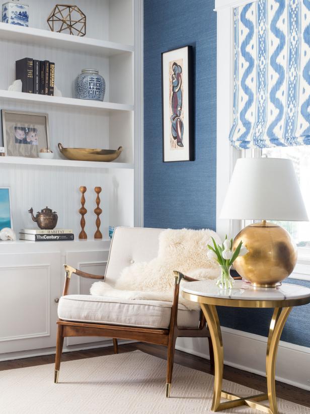 An elegant midcentury reading corner decorated with a white chair, blue grass cloth wallpaper and brass accents