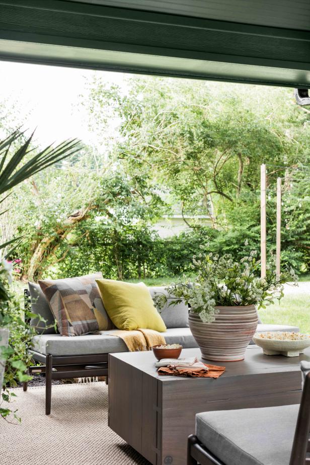Layers of pillows in tones of blue and yellow soften this outdoor seating area while blankets provide extra warmth on cool evenings. A large potted plant on the wooden coffee table adds a natural element to the outdoor living space.
