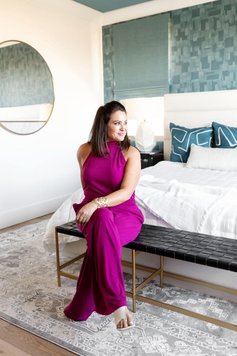 Woman with magenta jumpsuit sitting on white bed in teal bedroom.