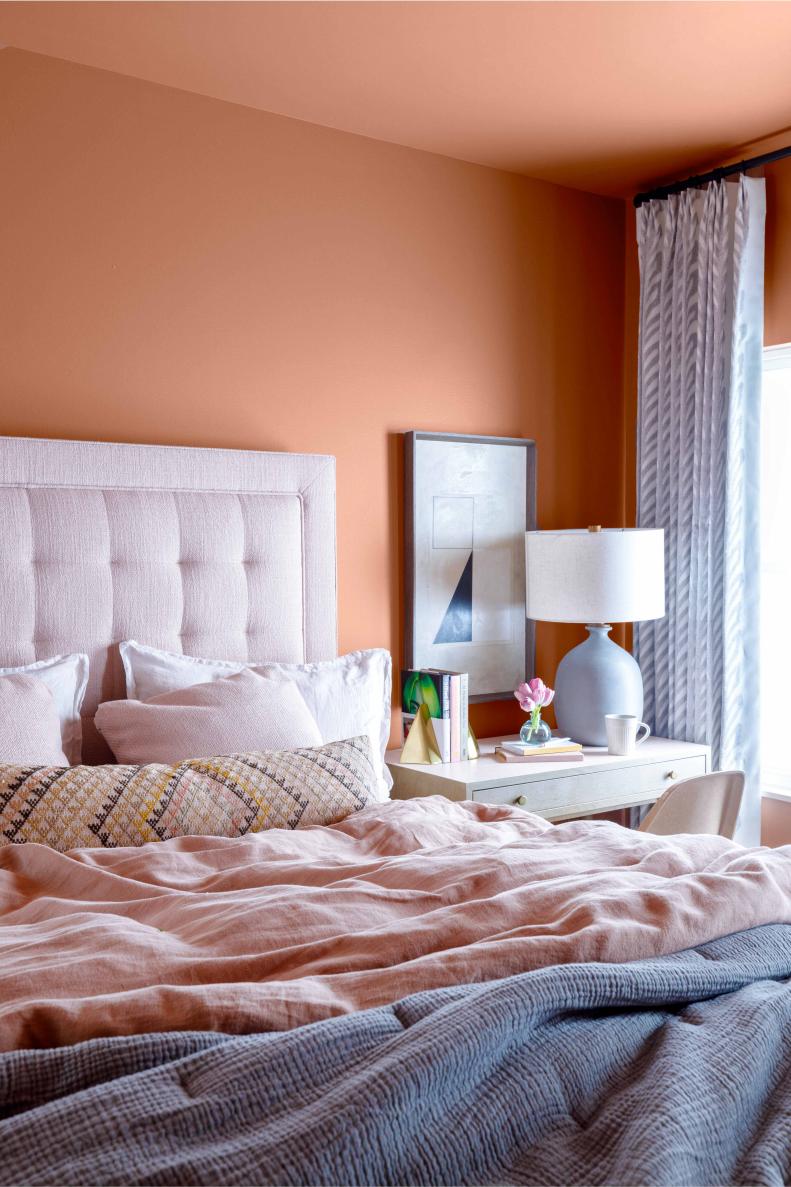 The pink bedding and upholstered headboard echo the warm tones found on the walls for a soft romantic feel. The side table lamp and drapes add texture in soothing gray for a relaxed vibe.