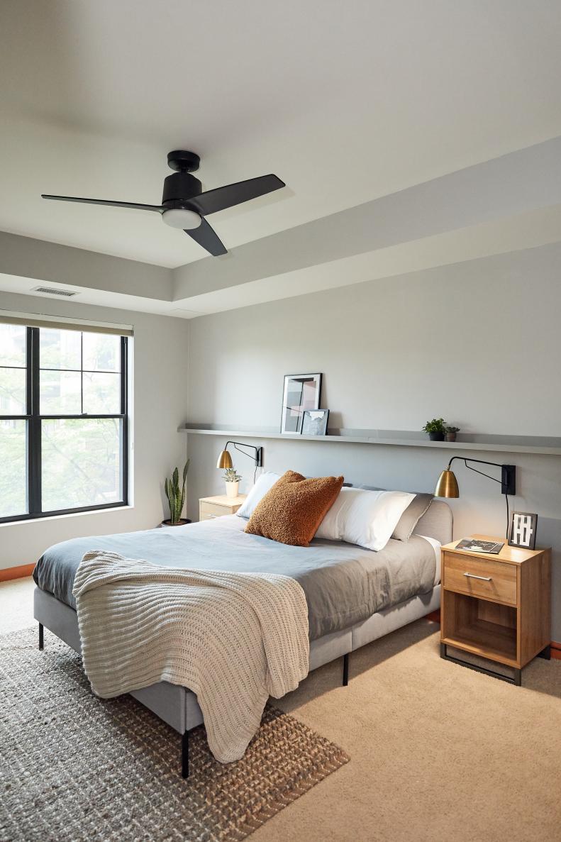 A modern gray bedroom decorated in minimalist style featuring a picture rail, art, a modern bed, ceiling fan, nightstands, wall sconces and a rug.
