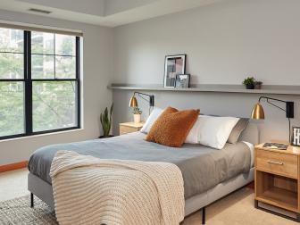 A modern bedroom with an upholstered bed, lamp, window shades, shelf for artwork and large window.