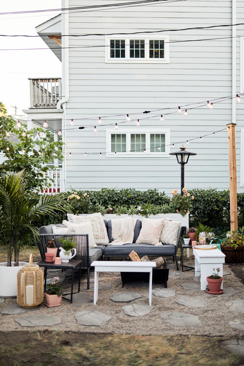 A paver and gravel patio anchors this outdoor space, framed by string lights, while an outdoor sofa and upcycled benches offer seating around a fire pit.