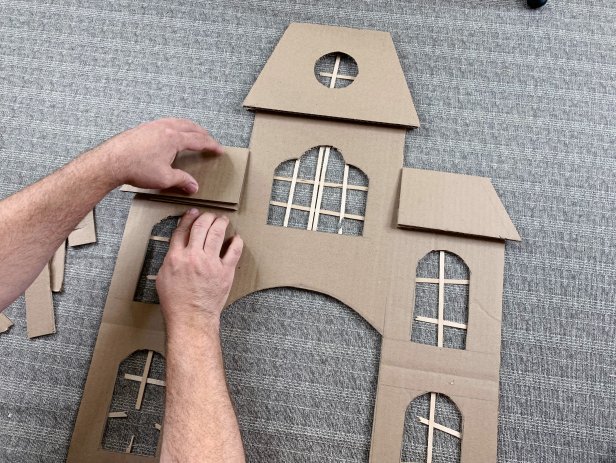 Make the house more dimensional by adding cardboard details. Cut pieces to match the size of the roof that is drawn on the facade. Create a slanted look by gluing small scrap strips of cardboard towards the bottom of the roof and glue into place. Rough up the shutter pieces by cutting cracks and hot gluing them slanted to the outside of each window.