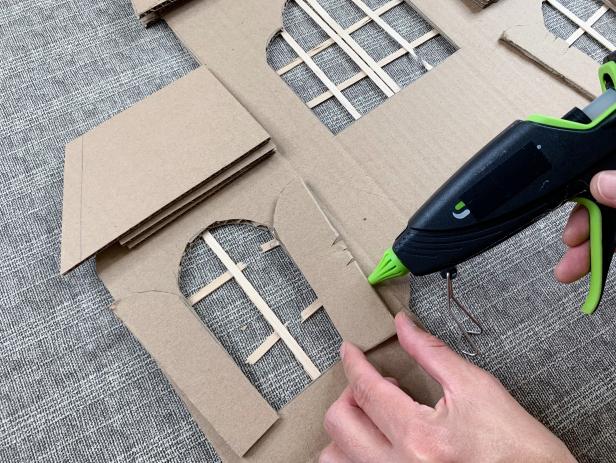 Make the house more dimensional by adding cardboard details. Cut pieces to match the size of the roof that is drawn on the facade. Create a slanted look by gluing small scrap strips of cardboard towards the bottom of the roof and glue into place. Rough up the shutter pieces by cutting cracks and hot gluing them slanted to the outside of each window.