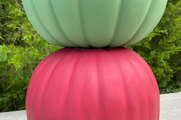 First, take a large nursery planter and turn it over; the planter bottom should be facing up. Now add a generous layer of super glue to the bottom of the largest, rust red, pumpkin. Move quickly to place the green pumpkin on top of the super glue layer. Press down on the green pumpkin to create a firm seal. Next, add a generous layer of super glue to the top of the green pumpkin. Move quickly to add the mauve pumpkin on top of the super glue layer. Press firmly to seal.