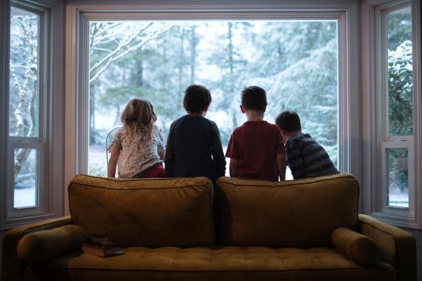 A group of elementary aged kids sit on the back of their living room couch, watching the winter snowflakes fall on their yard and trees.  Washington State, USA.