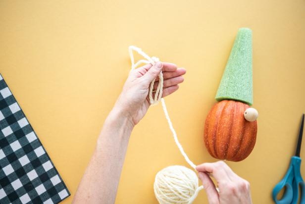 Yarn eyebrows for the no-carve pumpkin gnome project are made by loosely winding yarn around four fingers into a thick loop before securing the bundle with a yarn tie in the center and on each side. Hot glue secures the eyebrows to the gnome just above the wooden bead.