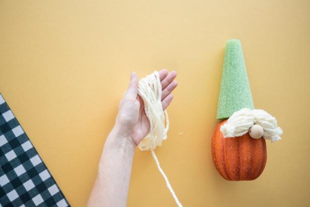 The no-carve pumpkin gnome's bushy beard is made by looping a large bundle of yarn around your hand until it's a large bundle that is secured by tying it off at the top and the bottom. The beard is glued to the pumpkin just below the wooden nose. The look can be customized with different colors of yarn.