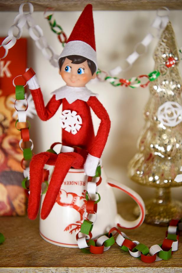 A toy elf sits on a coffee cup holding the tail of a paper chain.