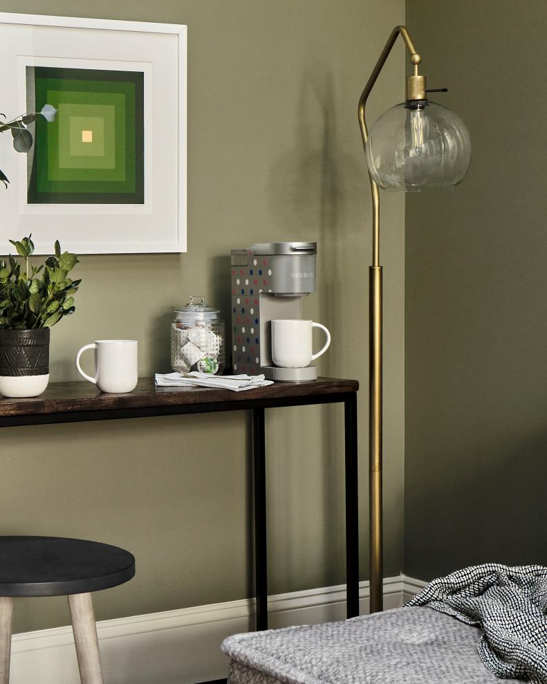 A modern console table includes space for a coffee station complete with beverage pods and a single serve coffee machine. A modern brass light with an exposed bulb adds a dash of metallic sparkle and design forward lighting.