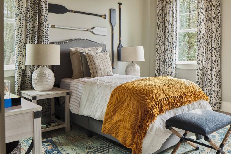 The guest room at HGTV Smart Home 2022 soothes with a mix of warm accents and soft sea-inspired details. The twin bed keeps a small footprint and is layered with cozy linens that add warmth to the space while the soft gray walls keep the room feeling calm and neutral.