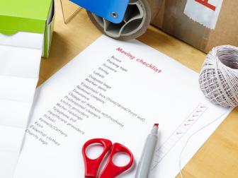 A house moving checklist on a table, surrounded by labels, packaging tape roller, scissors, red marker pen, a ball of string and a sealed box.  Some of the checkboxes have been ticked in red.