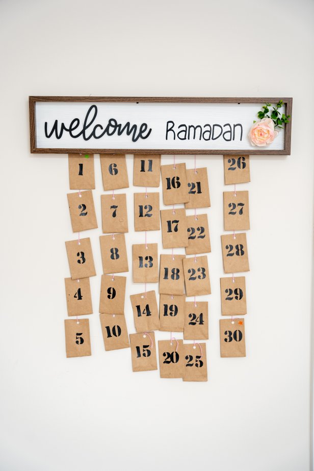 Areeba Adnan shares how to make an easy advent calendar to celebrate Ramadan. To make you'll need a welcome sign to upcycle, white and black paint, a low-temp hot glue gun, string, envelopes, stamps and faux florals for decoration.