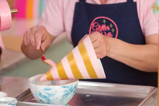 We decided to add pink glass glitter to the pink stripes. To add glitter, start by setting up a tray lined with parchment paper to catch the loose glitter. Paint a layer of tacky glue onto the desired areas (Image 1); a thick tacky glue helps embed the glitter into the glue and prevent fallout on the finished hat better than other glues. With the glue applied, add the glitter (Image 2). We put our glitter in a bowl and applied it with a spoon, then used a finger to tamp the glitter down. Avoid putting glitter or other ornate decoration near the bottom inch of the hat; this is where we'll need to cut the holes to tie on the hat's ribbon.