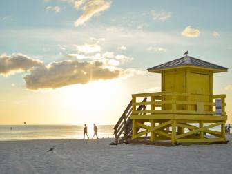 A couple walks past a yellow lifeguard station on the beach at sunset.