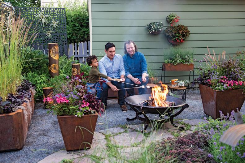 A small backyard space with a portable fire pit, wall baskets, container plants, decorative metal lanterns and two men and a boy roasting marshmallows.