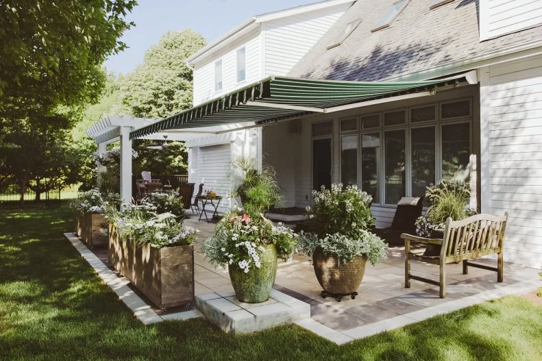 Throw Shade on Your Backyard A retractable awning and an arbor in a backyard with pots of flowers and foliage nearby.