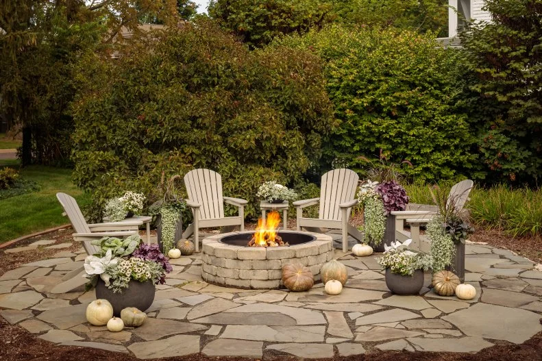 Add a Custom-Built Fire Pit A backyard patio made of pavers with a fire pit, Adirondack chairs, side tables, plants and ornamental pumpkins and gourds.