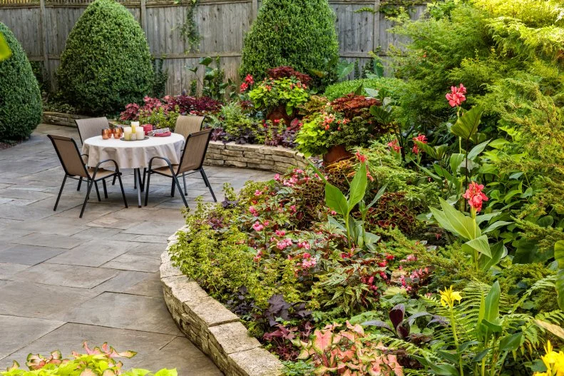 Shady Garden Backyard Landscaping A patio with a table and chairs enclosed by a curving border of pavers with plants.