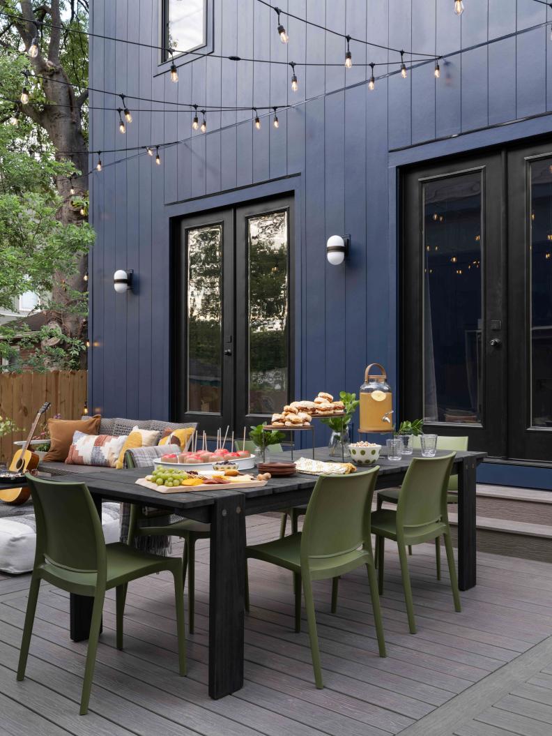 A large, black modern table with modern, green outdoor chairs anchors the backyard deck space. Globe lights strung from the house above the dining area add charm and light for evening gatherings.
