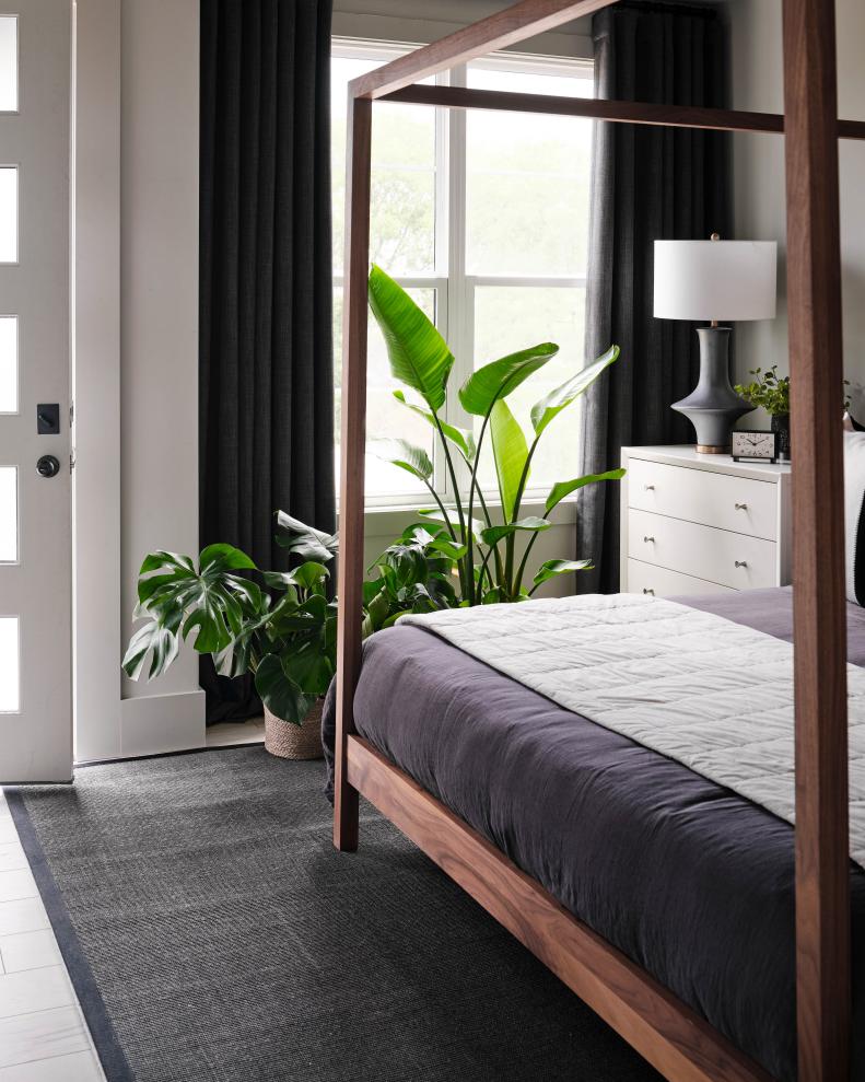 A dark gray rug beneath the bed anchors the sleeping area of the main bedroom and plays off the deep slate tones of the drapes. A series of potted plants beneath the window add life and greenery to the space for a lush, relaxed feel.