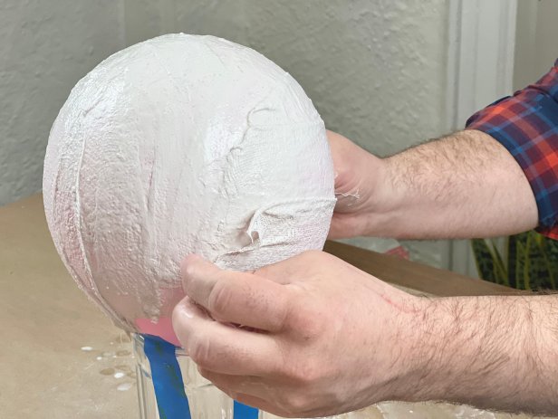 Work with one baddage at a time. Dip the bandage into the water for a few seconds and squeeze out any excess water with two fingers. Starting from the top. begin to layer the bandages onto the balloon. Once place on the balloon, rub your pointer finger in a swirling motion on top of the bandage. Doing this will help seal any holes that form and will also help keep the bandages down. Continue to add bandages around the balloon, leaving a hole where the balloon meets the cup. Tip: Try not to overlap the bandages too much or the egg shape will look lumpy.