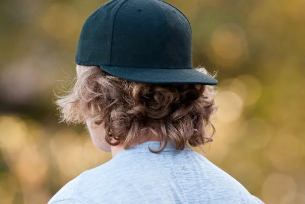 Teenage boy with long hair looking away in a black baseball hat.
