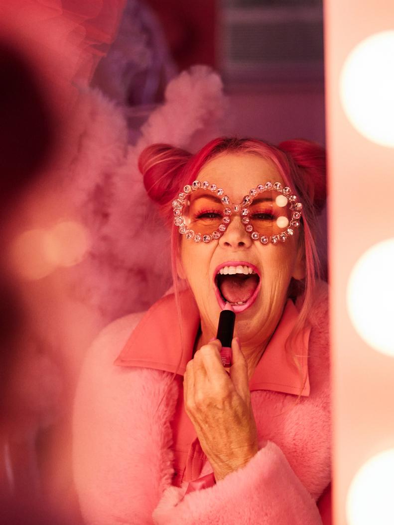 A woman applies pink lipstick while looking in a mirror.