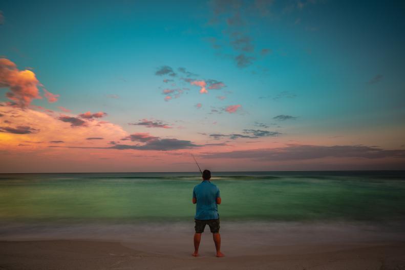 A man who is fishing, silhouetted against the sunset on a Gulf Coast beach.