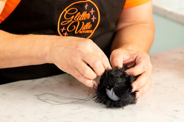 Step 7 of the low-sew spider hand puppet is creating the head by wrapping a piece of fur fabric around a styrofoam ball and securing it with a straight pin before sewing the sides shut with a running stitch.