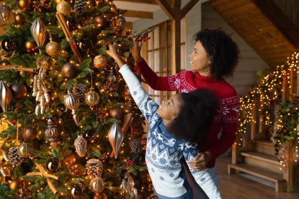 Happy African American woman with little daughter decorating Christmas tree with balls and baubles, smiling mother and adorable child girl, family wearing sweaters preparing home for New Year party