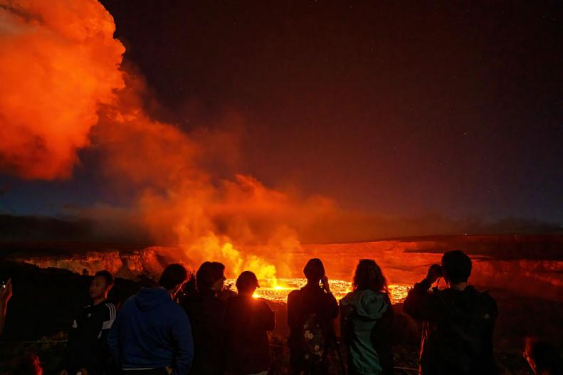 An aerial, nighttime view of a volcanic eruption in Hawaii with people watching the lava and smoke.