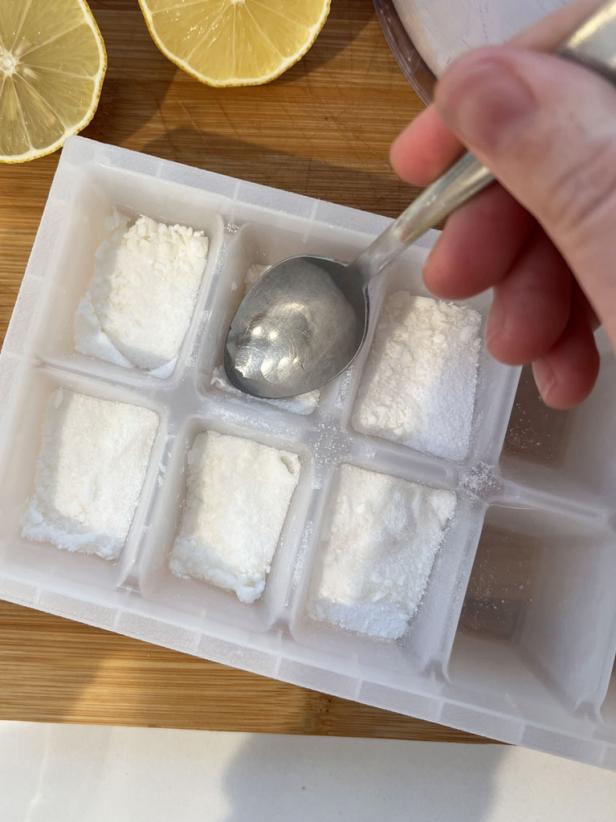 Using a spoon to press dishwasher detergent into ice cube trays with lemon juice to set.
