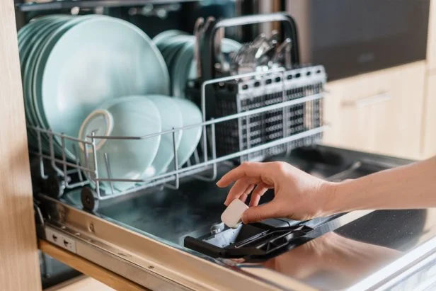 Close up view of female hand inserting dishwasher tablet into open built-in dishwasher machine with utensils inside in modern home kitchen. Household, housekeeping, domestic concept