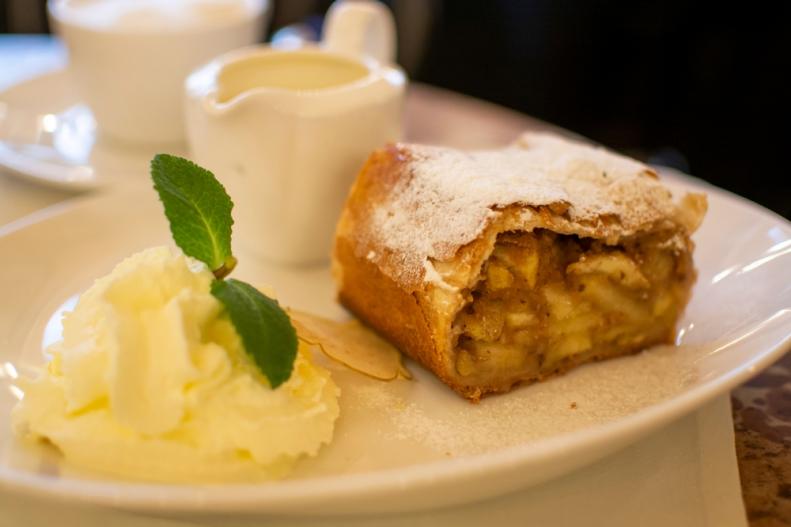 A close-up of a piece of Austrian apple strudel with whippen cream and hot vanilla sauce in a small pitcher on a tabletop