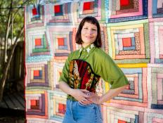 A woman stands in front of a log cabin quilt top hanging up outside.