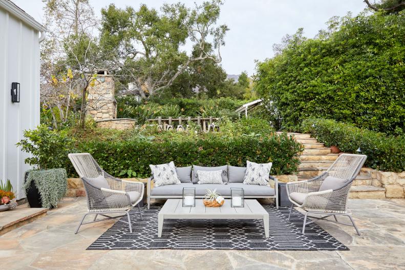 A patio with two chairs, a sofa, and an outdoor rug surrounded by overgrown shrubs and other plants.