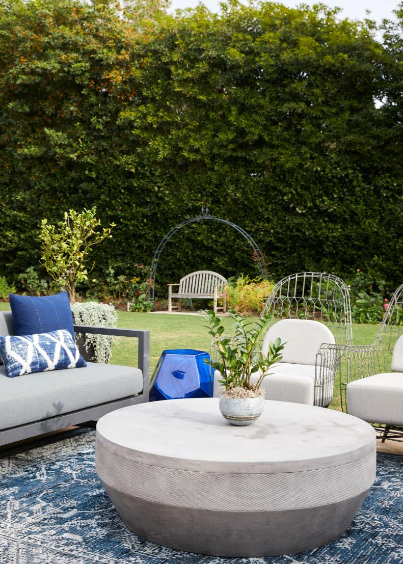 An outdoor living space featuring a round coffee table, sofa, chairs and a faceted accent table backed by tall trees and shrubs. 