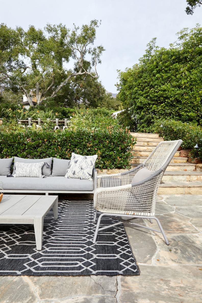 A sofa, chair and outdoor rug on a patio with shrubs and trees surrounding it.