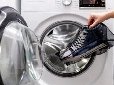 Woman putting blue sneakers in mesh laundry bag into washing machine, close up. Washing dirty sport shoes. Footwear care