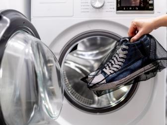 Woman putting blue sneakers in mesh laundry bag into washing machine, close up. Washing dirty sport shoes. Footwear care
