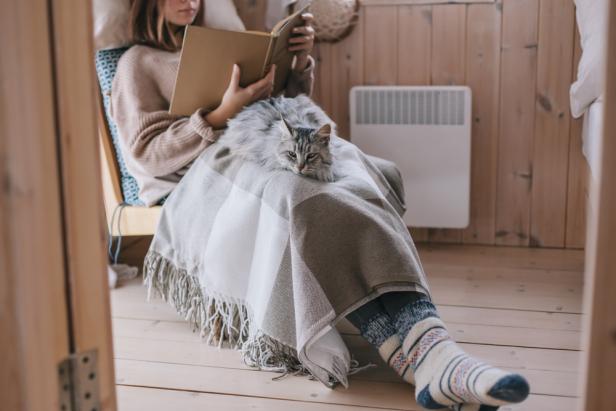 Young girl in sweater and warm socks relaxing on chair with cat and reading book in log cabin near electric heater