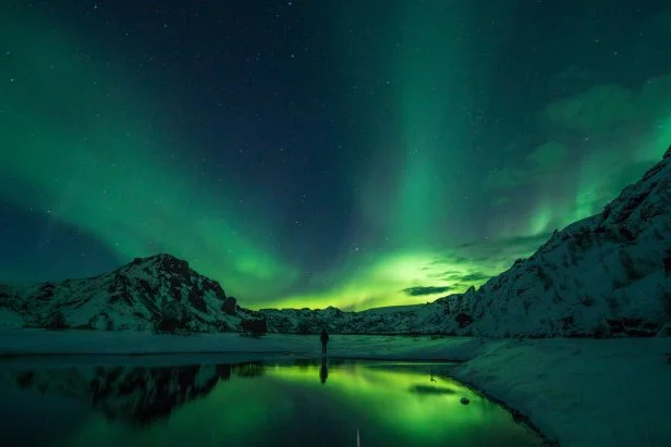 Someone standing in the mountains in Iceland with the Northern Lights shining overhead.