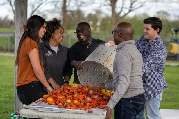 People standing around a table and dumping out a pot of boiled crawfish and other Cajun foods.