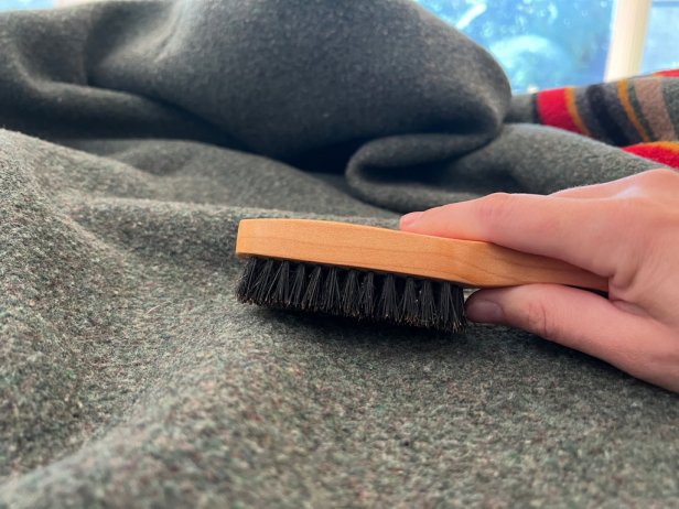 A soft-bristled hair brush loosens dust on a wool blanket.