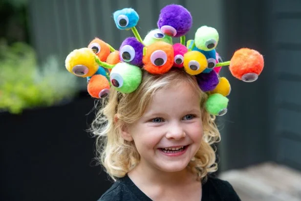 Girl wearing a colorful headband covered with pom-poms and google eyes.