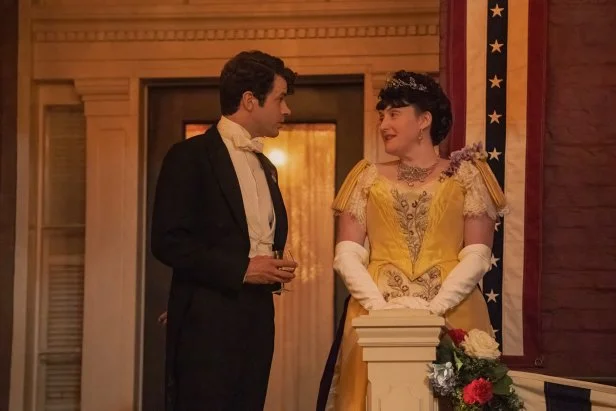 Man in tux and tails speaking with woman in yellow Victorian dress. 